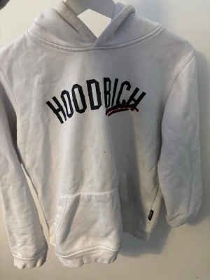 Vit hoodie från Hoodrich - Säljer en vit hoodie från Hoodrich med svart och röd text på bröstet och tryck på ryggen. Hoodien har en klassisk huva och en stor magficka framtill. Perfekt för dig som gillar streetwear och vill ha en clean look.
