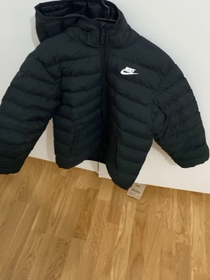 Svart pufferjacka från Nike - Snygg svart pufferjacka från Nike med huva och dragkedja framtill. Jackan har ett vitt Nike-logga på bröstet och är vadderad för extra värme. Perfekt för kalla dagar och enkel att matcha med olika outfits.