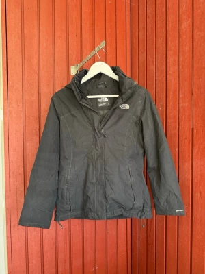 Svart vindjacka från The North Face - Svart vindjacka från The North Face med huva och dragkedja framtill. Jackan har två sidofickor med dragkedja och justerbar nederkant. Tillverkad i lätt och slitstarkt material, perfekt för blåsiga dagar. Klassisk logga på bröstet och ärmen.