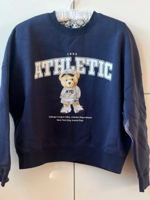 Mörkblå sweatshirt med björntryck - Cool mörkblå sweatshirt med stort ATHLETIC-tryck och en söt björn i streetwear på framsidan. Tröjan har rund hals, ribbade muddar och är gjord i mjukt bomullsmaterial. Perfekt för dig som gillar sportig och avslappnad stil.