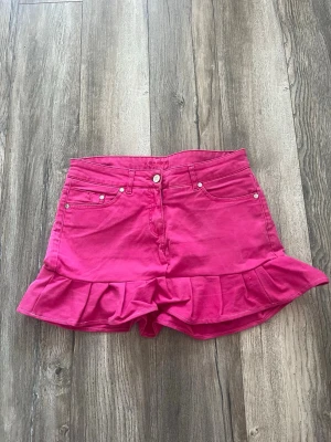 Rosa volangkjol med jeanslook - Snygg kort rosa kjol med volangkant och klassiska jeansdetaljer som fickor fram och bak samt bälteshällor. Kjolen har en feminin och lekfull stil, perfekt för dig som vill sticka ut med färg. Stängs med knapp och dragkedja framtill. (S/xs)