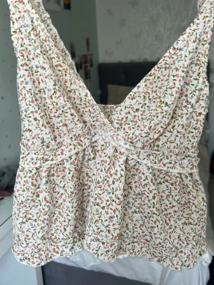 Blommig linnetopp med volang - Supersöt linnetopp med blommigt mönster i vitt, rosa och grönt. Toppen har volangdetaljer, v-ringning och smala axelband. Perfekt för varma dagar och ger en romantisk vibe till din outfit.