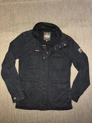 Mörkblå fieldjacket från Superdry - Snygg mörkblå jacka från Superdry med flera fickor framtill, ståkrage och detaljer som knappar och dragkedja. Jackan har patch på ärmen och Superdry-logga på bröstet. Perfekt för dig som gillar en stilren och funktionell look.