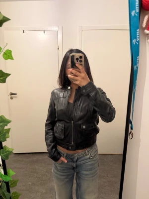 Svart skinnjacka från Stockh lm - Snygg svart skinnjacka från Stockh lm med två framfickor med lock och knapp, ståkrage och dragkedja framtill. Kortare modell som ger en cool och modern look. Perfekt till jeans eller kjol för en trendig outfit.