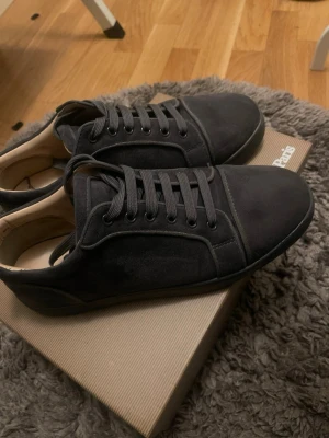 Gråa sneakers från Christian Louboutin - Stilrena gråa sneakers från Christian Louboutin i mocka med klassisk röd sula och svarta skosnören. Låg modell med diskreta sömmar och exklusiv känsla. Perfekta för dig som vill ha något lyxigt och tidlöst i garderoben.