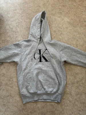 Grå hoodie från Calvin Klein Jeans - Snygg grå hoodie från Calvin Klein Jeans med stor CK-logga i svart på bröstet. Klassisk känguruficka framtill och justerbar huva med dragsko. Tillverkad i mjukt sweatshirtmaterial som är perfekt för en chill och avslappnad stil. Finns ingen storlek men skulle gissa på xs ungefär