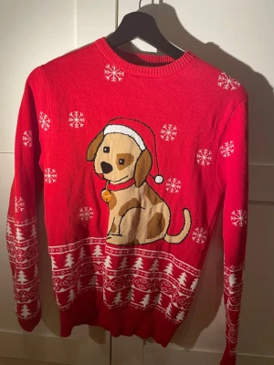 Röd jultröja med hundmotiv - Röd stickad jultröja från Christmas Sweats med gulligt hundmotiv i brunt och vitt, snöflingor och julgransmönster. Tröjan har rund hals och långa ärmar. Perfekt för julens alla tillfällen och för dig som gillar söta motiv.