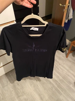 Svart Stone Island t-shirt - Snygg svart t-shirt från Stone Island med diskret logga broderad på bröstet och klassisk patch på ärmen. T-shirten har rund hals och korta ärmar, perfekt för en clean och stilren look. Tillverkad i mjuk bomull för skön komfort.