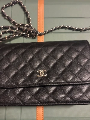 Svart Chanel kuvertväska i läder - Säljer en klassisk svart kuvertväska från Chanel i quiltat läder med silverfärgad kedja och ikonisk CC-logga framtill. Väskan har magnetlås, röd innerficka med dragkedja och guldfärgad Chanel-stämpel på insidan. Perfekt för dig som gillar tidlös och lyxig design.