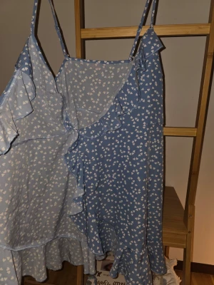 Blå omlottklänning med volanger - Söt blå omlottklänning med vita hjärtan, tunna axelband och volangdetaljer längs kanterna. Klänningen har knyt i midjan och är i en kort modell, perfekt för sommarens varma dagar.