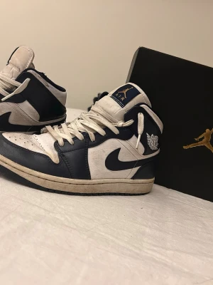Air Jordan 1 - Säljer ett par Air Jordan 1 Retro High OG i marinblått och vitt. Klassisk high-top siluett med vita skosnören, läderpaneler och ikoniska Air Jordan-loggan på sidan. Snygg kontrast mellan färgerna och en tidlös design som alltid är aktuell.
