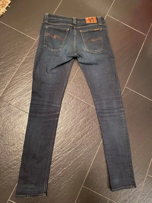 Mörkblåa Nudie Jeans -  par mörkblåa jeans från Nudie Jeans med klassiska bakfickor och kontrastsömmar. Jeansen har en smal passform. Snygg detalj med Nudie-loggan på bakfickan och läderpatch i midjan.