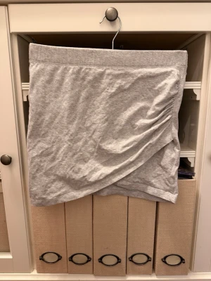 Grå omlottkjol i bomull - Snygg grå kortkjol i mjuk bomull med omlottdesign och lätt rynkning på sidan. Perfekt för en avslappnad och trendig look. Enkel att matcha med olika toppar och sneakers.