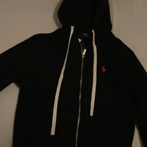 Svart hoodie från Polo Ralph Lauren - Snygg svart hoodie med dragkedja från Polo Ralph Lauren. Tröjan har vit dragsko i huvan och den klassiska röda loggan broderad på bröstet. Perfekt för en avslappnad och stilren look.