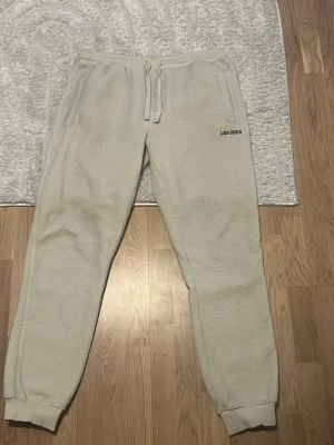 Beiga fleecebyxor från Les Deux - Mysiga beiga mjukisbyxor från Les Deux i fluffigt fleecematerial. Byxorna har resår i midjan med dragsko, muddar vid bensluten och sidofickor. Perfekta för chill dagar hemma eller när du vill vara extra bekväm.