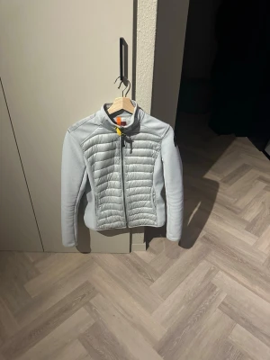Ljusgrå vindjacka från Parajumpers - Snygg ljusgrå vindjacka från Parajumpers med quiltade partier fram och bak, hög krage och dragkedja. Jackan har sidofickor med dragkedja och en extra ficka bak. Materialmix med slät och vadderad yta samt diskret logga på ärmen.