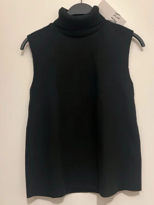 Svart ärmlös polotröja från Zara  - Stilren svart ärmlös polotröja från Zara med hög ribbad krage. Tröjan har rak passform och är tillverkad i ett mjukt, stickat material som känns skönt mot huden. Perfekt att matcha med skjorta eller t-shirt under för en trendig lager-på-lager-look.