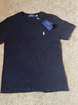 Mörkblå t-shirt från Polo Ralph Lauren - Snygg mörkblå t-shirt från Polo Ralph Lauren med klassisk passform och korta ärmar. T-shirten har en liten vit broderad logga på bröstet och är tillverkad i mjuk bomull. Perfekt basplagg för en clean och stilren look.