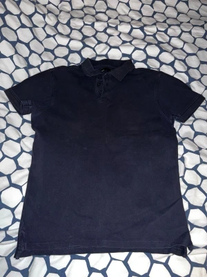 Mörkblå pikétröja med krage - Klassisk mörkblå pikétröja med krage och knappar framtill. Tröjan har korta ärmar och en enkel, stilren design som passar till det mesta. Perfekt för dig som gillar en clean och tidlös look.