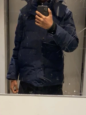 Mörkblå canada goose pufferjacka med huva - Snygg mörkblå pufferjacka med hög krage och huva. Jackan har en boxig passform, stora paneler och stängs med både dragkedja och knappar. Perfekt för kalla dagar och ger en modern streetwear-look.