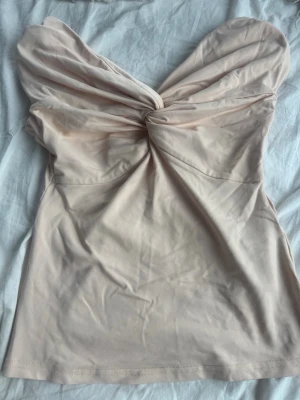 Beige axelbandslös omlottopp - Snygg beige axelbandslös topp med omlottdetalj och twist framtill. Toppen har en croppad känsla och är tillverkad i mjukt stretchigt material som formar sig efter kroppen. Perfekt för dig som vill ha en stilren och trendig look.