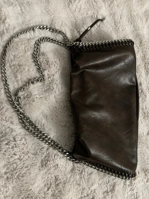 Mörkbrun kedjeväska i fejkläder ginatricot - Snygg mörkbrun handväska i fejkläder med silverfärgad kedja som axelrem och detalj runt kanten. Väskan har en mjuk, slouchy form och stängs med magnetknapp. Perfekt för dig som gillar en cool och trendig look.