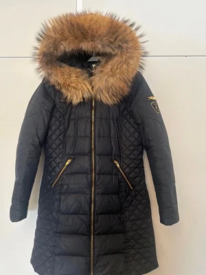 Svart dunjacka Rockandblue med pälshuva - Snygg svart lång dunjacka från Rockandblue med stor pälskantad huva och guldfärgade detaljer. Jackan har två snedställda fickor med dragkedja och Rockandblue-märke på ärmen. Quiltade partier och figurnära passform ger en modern look.