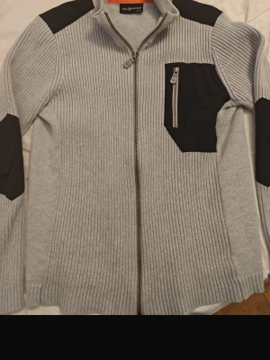 Ljusgrå stickad ziptröja sail racing  - Säljer en ljusgrå stickad ziptröja sail racing  med svarta detaljer på axlar, armbågar och bröstficka. Tröjan har ribbad struktur, hög krage och en praktisk bröstficka med dragkedja. Perfekt för dig som gillar stilren och sportig look.