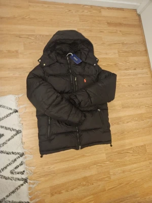 Rahlp lauren puffer - Snygg svart pufferjacka från Polo Ralph Lauren med huva och dragkedja framtill. Jackan har två sidofickor med dragkedja och den klassiska röda loggan broderad på bröstet. Perfekt för kalla vinterdagar och riktigt skön att bära.