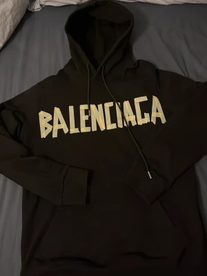 Svart Balenciaga hoodie - Säljer en svart hoodie från Balenciaga med stort gult tryck på bröstet och ryggen. På framsidan står det 'BALENCIAGA'. Hoodien har huva med dragsko och ribbade muddar. Perfekt för dig som gillar streetwear och statement-plagg.Passar även M. Kan gå ner till 1900-2000 vid snabb affär. 