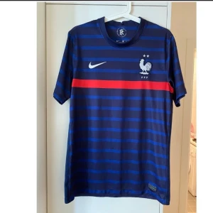 Frankrike Nike matchtröja blå med keps  - Snygg blå matchtröja från Nike med Frankrikes landslagslogga och två stjärnor på bröstet. Tröjan har horisontella mörkblå ränder, en röd rand över bröstet och vit Nike-logga. Tillverkad i lätt och ventilerande material, perfekt för fotboll eller träning.