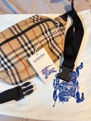 Beige bälte-väska från Burberry - Säljer en beige bälte-väska från Burberry med det klassiska rutmönstret i svart, vitt och rött. Väskan har ett justerbart svart band med spänne och dragkedja upptill. Perfekt för att bära det viktigaste och ger en stilren touch till din outfit.
