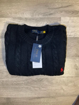 Svart kabelstickad tröja Polo Ralph Lauren - Kabelstickad svart tröja från Polo Ralph Lauren med rund halsringning och ribbade muddar. Tröjan har det klassiska röda Polo-logot på bröstet och är tillverkad i mjukt material. Perfekt för dig som vill ha en stilren och tidlös look.