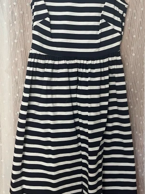 Randig axelbandslös klänning från Lindex - Snygg axelbandslös klänning med marinblå och vita horisontella ränder. Klänningen har en utställd kjol och markerad midja för en feminin siluett. Perfekt för sommarens alla tillfällen och enkel att styla med accessoarer. Vit och marinblå-randig. Stl. 36