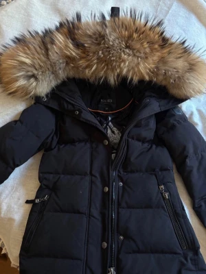 Svart parkas med pälskant från Holies - Snygg svart parkas från Holies med stor, fluffig pälskant i bruna toner på huvan. Jackan har dragkedja och knappar framtill, två snedställda fickor med dragkedja och justerbar midja. Perfekt för kalla dagar och riktigt varm.