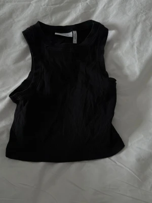 Svart ribbad linnetopp från Weekday - Säljer en svart ribbad linnetopp från Weekday i storlek S. Toppen är ärmlös med rund halsringning och har en croppad passform. Perfekt att styla till jeans eller kjol för en clean och enkel look.