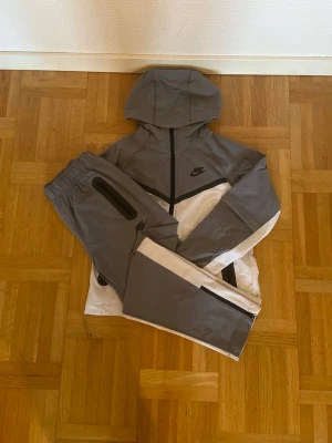 Nike grå och vit träningsdress - Nike träningsdress i grått och vitt med svarta detaljer. Setet består av en zip-hoodie med huva och matchande byxor med dragkedja vid benslut och fickor. Tillverkad i lätt och funktionellt material, perfekt för träning eller chill.