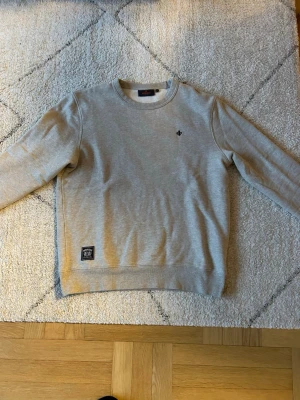 Grå sweatshirt från Morris - Snygg grå sweatshirt från Morris med rund halsringning och diskret logga broderad på bröstet. Tröjan har ribbade muddar vid ärmslut och nederkant samt en liten patch nertill. Perfekt till ett avslappnat streetwear-look. Sparsamt använd då det är en present och var snudd på för liten direkt. 