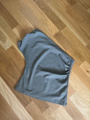 Grå oneshoulder topp - Snygg grå oneshoulder topp i mjukt bomullsmaterial. Toppen har en enkel och stilren design med en axel och lätt rynkning vid sidan för en cool look. Perfekt att matcha med jeans eller kjol.