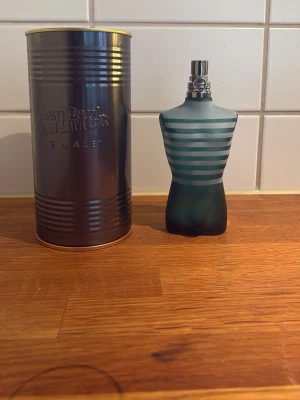 Jean Paul Gaultier Le Male parfym - Klassisk herrparfym från Jean Paul Gaultier, Le Male. Flaskan har en unik design i form av en manlig torso med blå och turkosa ränder. Kommer i en metallisk cylinderformad förpackning. Flaskan är av frostat glas och har silverfärgad spraytopp. 100 milliliter kvar
