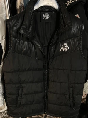 Svart dunväst från Kings Will Dream - Snygg svart dunväst från Kings Will Dream med broderad logga på bröstet. Västen har hög krage, dragkedja framtill och två fickor med dragkedja. Perfekt för lager-på-lager och streetwear-stil.