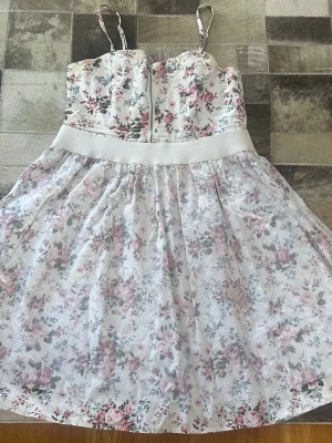 Blommig sommarklänning med smala band stl 170 - Söt kortklänning med smala axelband och blommigt mönster i rosa och lila toner på vit botten. Klänningen har en markerad midja med bred resår och lätt utställd kjol. Perfekt för varma dagar och har en romantisk känsla.