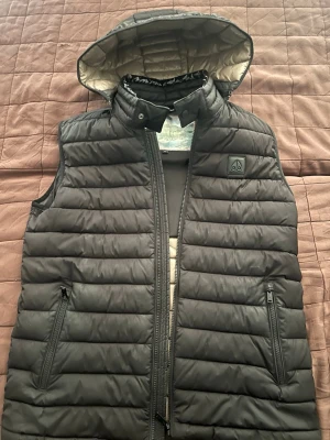 Svart dunväst från moose knuckles  - Snygg svart dunväst från moose knuckles med avtagbar huva och dragkedjor på fickorna. Västen har quiltad design och beige insida. Perfekt för lager-på-lager och håller dig varm under kyliga dagar. Klassisk logga på bröstet.