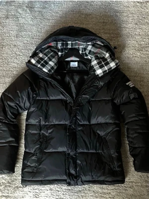 Svart dunjacka med huva från Moncler - Snygg svart dunjacka från Burberry med stor huva och rutigt innerfoder i svart, vitt och grått. Jackan har dragkedja och knappar framtill, två snedställda fickor med dragkedja och Burberry logga på ärmen. Perfekt för kalla vinterdagar.