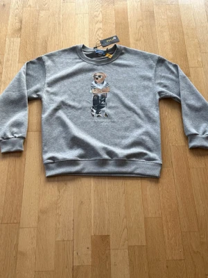 Grå sweatshirt från Polo Ralph Lauren - Snygg grå sweatshirt från Polo Ralph Lauren med ikoniskt björnmotiv framtill. Tröjan har rund hals, ribbade muddar och är tillverkad i mjuk bomull. Perfekt för en avslappnad och trendig look.