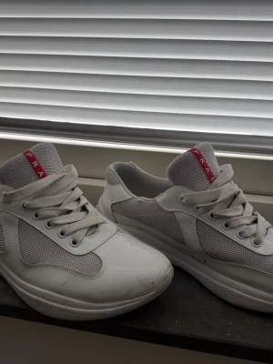 Vita sneakers från Prada - Boxen till skorna tillkommer. Snygga vita sneakers från Prada med meshdetaljer och klassisk röd logga på plösen. Skorna har vita snören och en chunky sula som ger en modern vibe. Perfekta för dig som vill ha en clean och stilren look med exklusiv känsla.