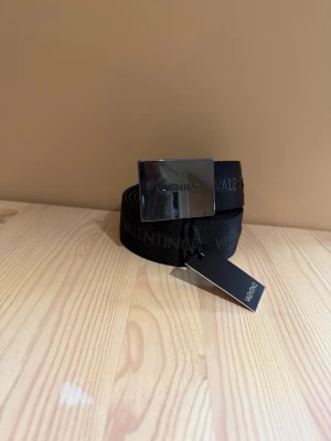 Svart Valentino bälte med silverdetalj - Snyggt svart bälte från Valentino med bred silverfärgad metallspänne där märket är ingraverat. Bältet har diskret Valentino-text längs bandet och passar perfekt till jeans eller byxor för en clean och modern look. 110-120cm