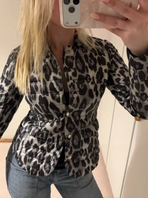 Leopardmönstrad pufferjacka - Säljer en snygg leopardmönstrad pufferjacka med tryckknappar framtill. Jackan har figurnära passform, hög krage och är tillverkad i ett glansigt material. Perfekt för dig som vill sticka ut i vinter och gillar djurmönster.