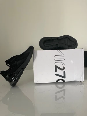 Nike Air Max 270 helsvarta sneakers - Nike Air Max 270 sneakers i helsvart med synlig Air-enhet i hälen. Ovandelen är i mesh och syntetmaterial för bra andningsförmåga och komfort. Klassisk snörning och modern, sportig design som passar till streetwear. Snygga detaljer och logga på sidan.