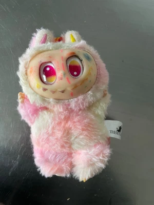 TheMonsters rosa/vit samlarfigur - Unik samlarfigur från TheMonsters med fluffig päls i rosa och vitt. Figuren har stora rosa ögon, färgglada detaljer i ansiktet och små öron på huvudet. Perfekt för dig som gillar udda och kreativa prydnader. Kan vara några streck med tuschpenna av ett barn 